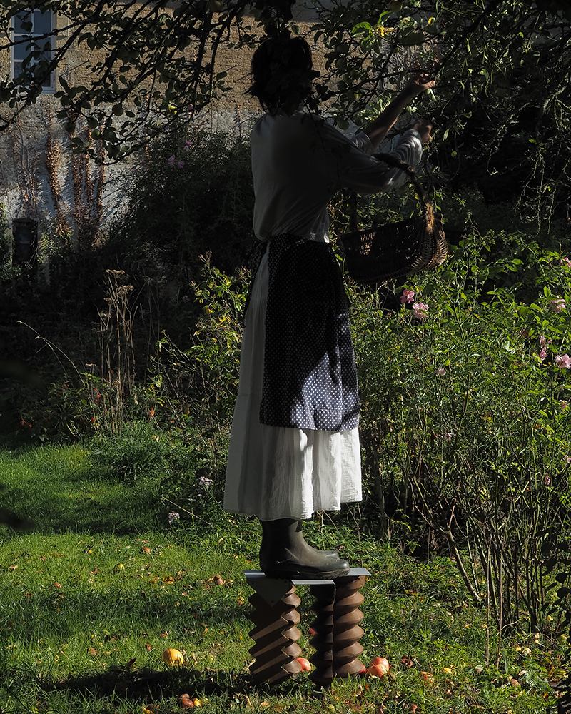 woman stands atop the wood and aluminum stool in a green garden