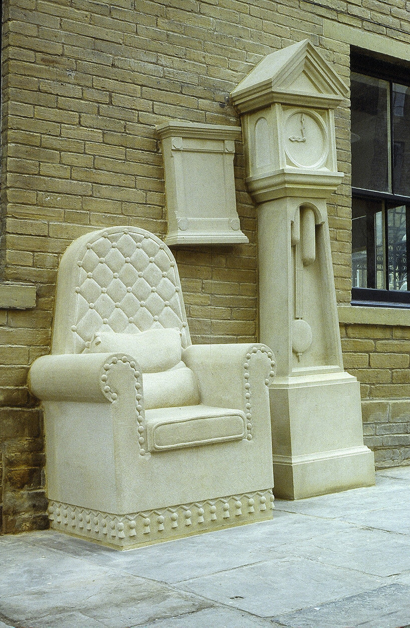 stone-carved grandad’s clock and chair by timothy shutter sit on a sloped street in england