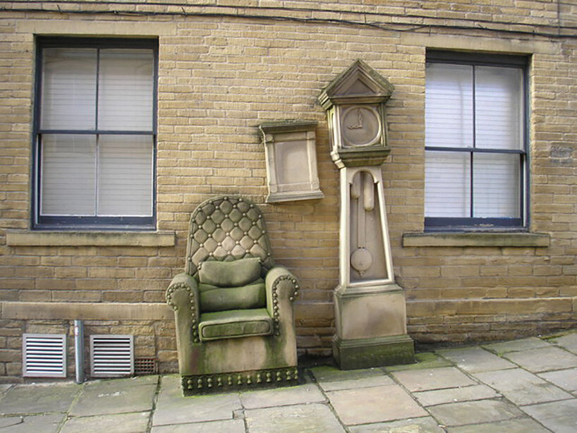 stone-carved grandad’s clock and chair by timothy shutter sit on a sloped street in england