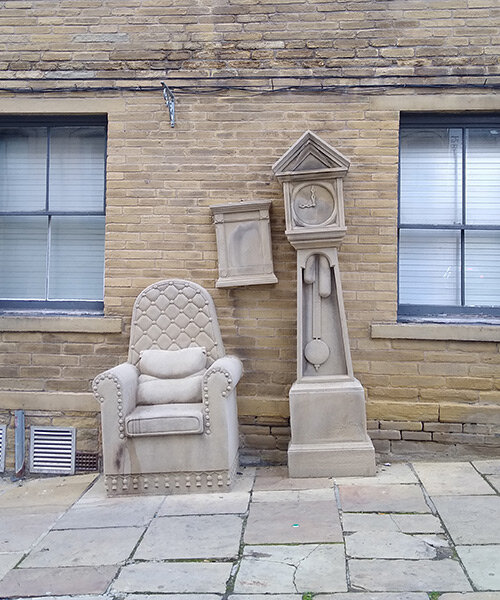 stone-carved grandad’s clock and chair by timothy shutter sit on a sloped street in england
