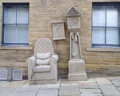 stone-carved grandad’s clock and chair by timothy shutter sit on a sloped street in england