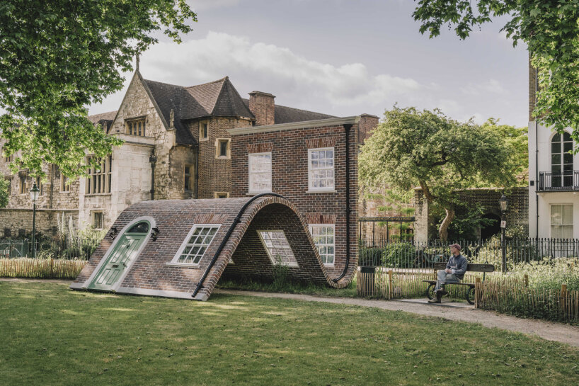 Alex Chinneck sitting next to his monumental brick facade sculpture named 'A week at the knees'