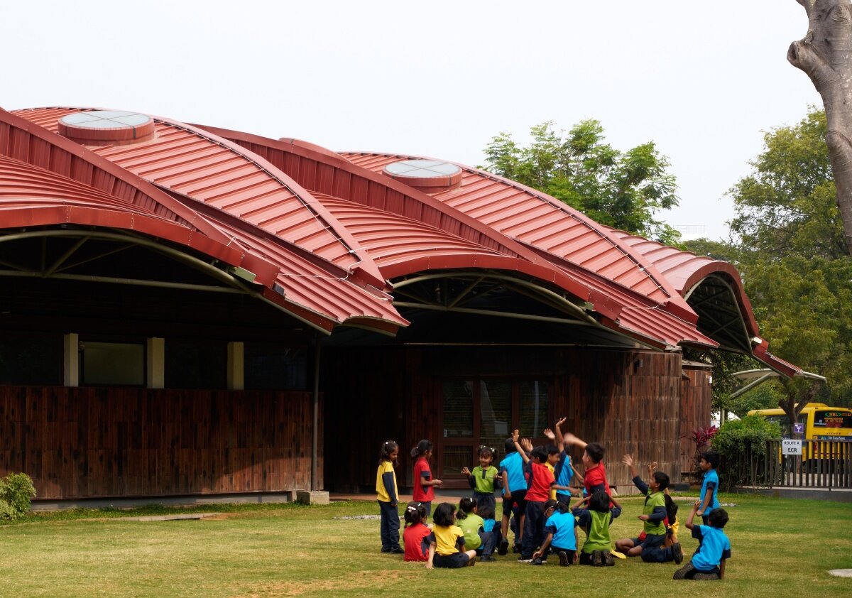 layered red roof sweeps over vastushilpa sangath’s tree-filled school in india