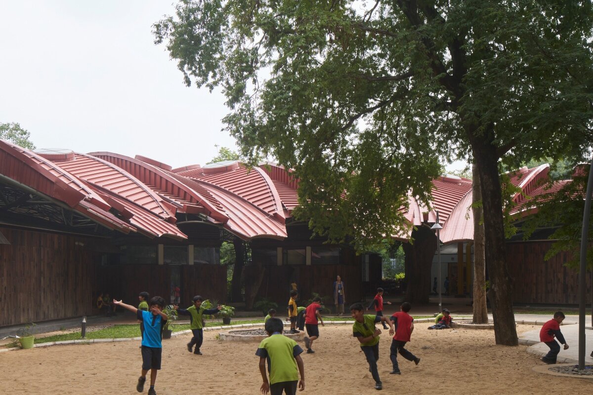layered red roof sweeps over vastushilpa sangath’s tree-filled school in india