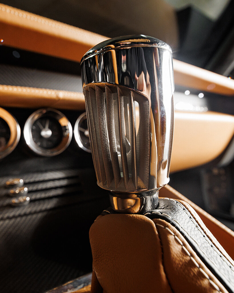 The image shows a close-up of a luxury car's gear shift knob, which is made of polished metal with vertical grooves and reflects its surroundings. The gear shift is mounted on a base wrapped in tan and black leather with visible stitching. In the background, part of the car's dashboard is visible, featuring analog gauges with chrome bezels and a section covered in tan leather, contributing to the overall high-end interior design.