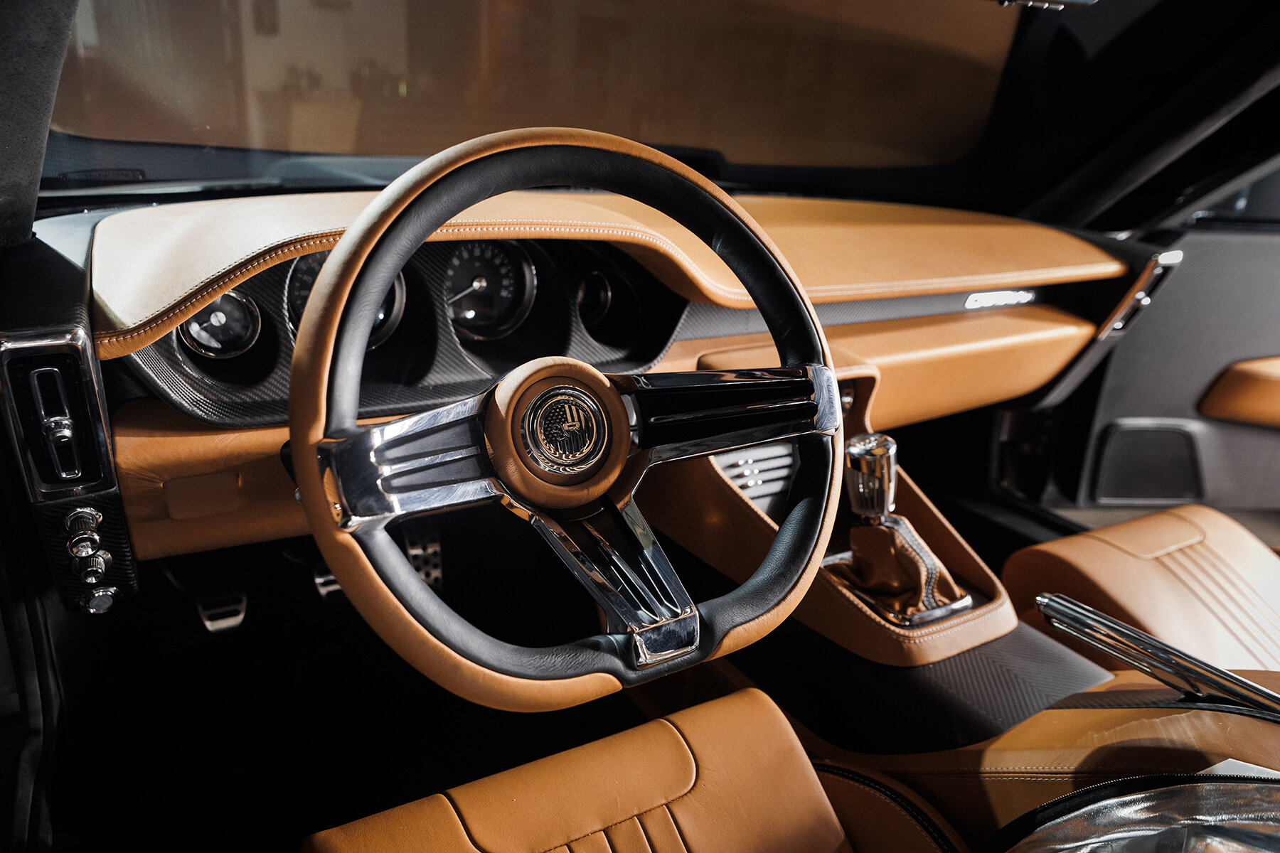 The image shows the interior of a car, focusing on the steering wheel and dashboard area. The steering wheel has a classic design with a polished metal three-spoke center wrapped in black and tan leather. The dashboard and seats are upholstered in tan leather with detailed stitching, and the gear shift is wrapped in matching tan leather. The overall interior has a vintage, luxurious look with a mix of modern touches such as the carbon fiber-style paneling around the gauges.