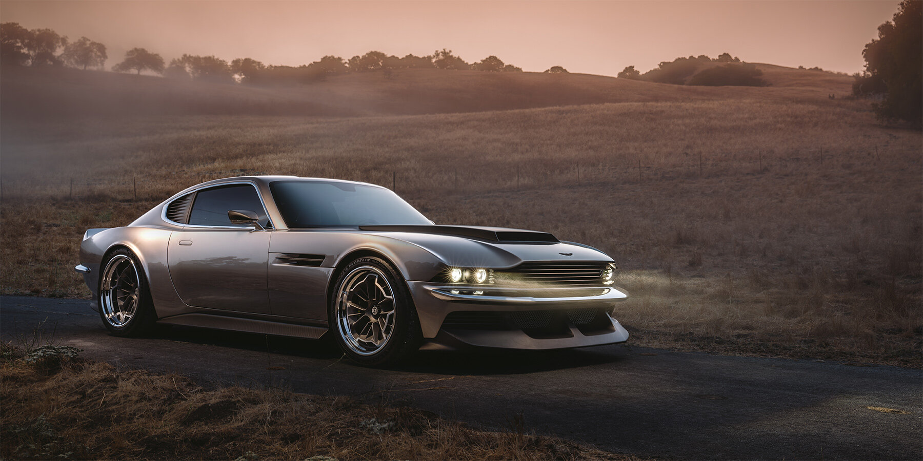 A sleek, metallic silver sports car is parked on a narrow paved road amidst a dry, grassy landscape with rolling hills in the background. The car has a low profile with wide, polished wheels and its headlights are on, casting beams of light that subtly illuminate the pavement ahead. The sky is tinged with soft, warm hues of sunset, creating a serene and slightly misty atmosphere around the scene.